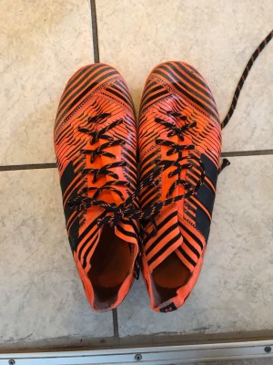 Adidas orangea fotbollsskor med mönster - Säljer ett par grymma Adidas fotbollsskor i orange med svarta ränder och grafiskt mönster. Skorna har snörning och låg profil, perfekta för snabba rörelser på planen. Tillverkade i syntetmaterial för bra grepp och känsla. Riktigt snygg design som sticker ut!