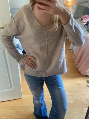 Beige v-ringad tröja - En beige v-ringad tröja med lång ärm och avslappnad passform. Tröjan har en dekorativ söm framtill och är gjord i ett mjukt, stickat material som känns skönt mot huden. Perfekt för dig som gillar en enkel och stilren look. 100% Wool