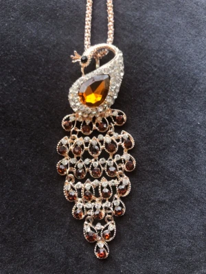 Långt statement-halsband med påfågel – Guldton & Strass (bärnstens bruna nyanser) - Säljer ett superfint och detaljrikt halsband med ett stort hänge format som en påfågel. Smycket är prytt med gnistrande stenar i klara och bärnstensfärgade/bruna nyanser. Viktigt att veta: Eftersom det är ett bijouteri med dekorativa stenar (ej äkta ädelstenar) så väger halsbandet nästan ingenting. Det är med andra ord väldigt bekvämt att bära trots sin storlek! • Skick: Mycket gott skick, inga stenar fattas. • Mått på hänge: Ca 10 cm långt och 3-4 cm brett. • Hela längden: Ca 46 cm 