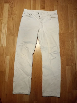 Vita raka jeans med knappar - Säljer ett par vita jeans med rak passform och knappgylf. Klassisk femficksmodell och bälteshällor. Jeansen har en enkel och clean look, perfekta till många olika stilar. Materialet känns som bomull med lite stretch.