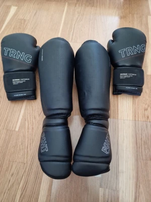 Svarta boxningshandskar och benskydd - Säljer ett set med svarta boxningshandskar och benskydd från Decathlon Outshock. Handskarna har TRNG-tryck och kardborrestängning, benskydden har FIGHT-tryck. Tillverkade i slitstarkt syntetmaterial, perfekta för kampsportsträning. Handskarna är storlek 14oz och benskydden är M