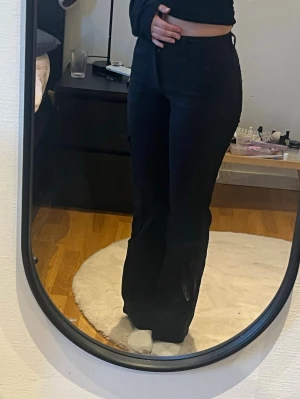 Svarta utsvängda jeans - Säljer ett par svarta jeans med hög midja och utsvängda ben. Jeansen har en stilren look med klassisk knapp- och dragkedjestängning framtill. Materialet är mjukt och följsamt, vilket gör dem riktigt bekväma och trendiga.