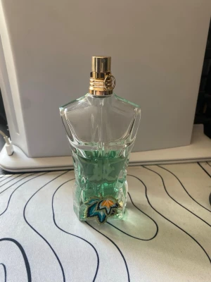 Jean Paul Gaultier Le Male parfym - Säljer en ikonisk Jean Paul Gaultier Le Male parfym i en unik flaska formad som en manlig torso. Flaskan är genomskinlig med turkosgrön vätska och har ett guldfärgat sprayhuvud. Perfekt för dig som gillar klassiska dofter och snygg design.