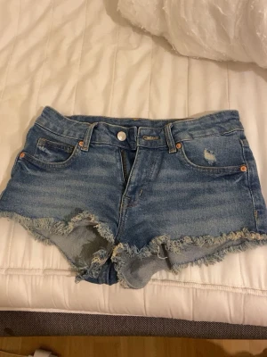 Blå jeansshorts med fransig kant - Snygga blå jeansshorts med fransig och sliten kant samt små slitningar framtill och baktill. Klassisk femficksmodell med knapp och dragkedja. Perfekta för sommaren och ger en cool, avslappnad look.
