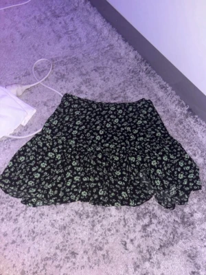 Svart blommig kortkjol med volang - Supersöt svart kortkjol med små gröna blommor och volangkant. Kjolen har en flowy passform och är perfekt för vår och sommar. Lätt att matcha med både t-shirt och stickad tröja.