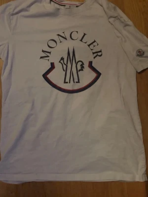 Vit Moncler t-shirt med logga - Vit t-shirt från Moncler med stor tryckt logga på bröstet och Moncler-märke på ärmen. Klassisk passform och rund halsringning. Perfekt för en stilren och trendig look.