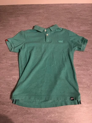 Grön pikétröja från Superdry - Snygg grön pikétröja från Superdry med klassisk krage och knappar framtill. Tröjan har korta ärmar och diskret broderad logga på bröstet. Perfekt för en avslappnad och stilren look.