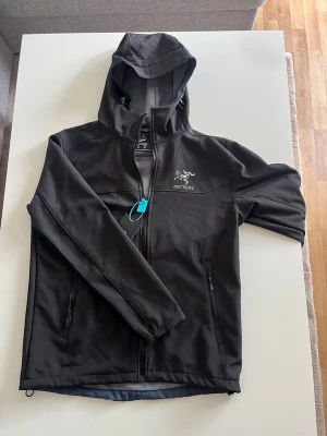 Svart vindjacka från Arc'teryx - Svart vindjacka från Arc'teryx med huva och dragkedja framtill. Aldrig använd. Storlek XL men sitter som L