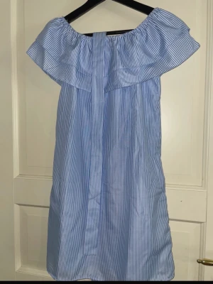 Blåvit off shoulder klänning - Snygg blå och vitrandig off shoulder-klänning med volang upptill. Klänningen har raka linjer och är tillverkad i ett lätt tyg, perfekt för varma dagar. Klassisk och fräsch look med smala vertikala ränder.