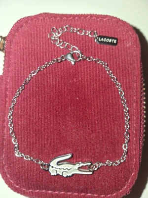 Silverfärgat armband från Lacoste - Snyggt och stilrent armband i silverfärg från Lacoste med den ikoniska krokodil-loggan. Kedjan är tunn och har ett justerbart spänne samt en liten bricka med Lacoste-gravyr. Perfekt accessoar för att lyfta din outfit.