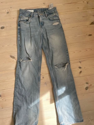 Ljusblå ripped jeans från Zara - Säljer ett par populära ljusblå jeans från Zara med vida ben, lågmidja och slitna detaljer vid knäna och fickorna. Jeansen har klassisk femficksmodell.. Perfekta för en avslappnad och trendig look.
