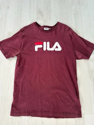 Vinröd FILA t-shirt med logga - Säljer en vinröd t-shirt från FILA med stor vit och röd logga på bröstet. Klassisk rund halsringning och korta ärmar. Tillverkad i mjuk bomull som är skön att bära. Perfekt för en avslappnad och sportig stil.