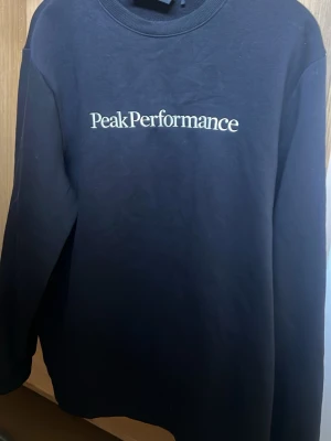 Svart Peak Performance sweatshirt - Svart sweatshirt från Peak Performance med vit logga tryckt på bröstet. Klassisk rund halsringning och långa ärmar. Perfekt för en avslappnad och stilren look.