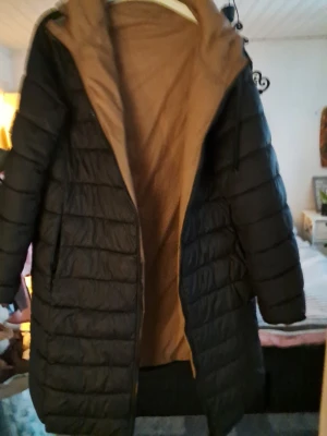 Svart lång pufferjacka med brun insida - Snygg svart pufferjacka med lång passform och brun insida. Jackan har dragkedja framtill, två sidofickor och en hög krage. Perfekt för kalla dagar när du vill hålla dig varm och stilren.