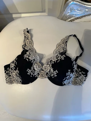 Svart spets-bh från Cream 85C - Snygg svart bh från Cream med vacker beige spetsdetalj över kuporna och axelbanden. Bh:n har bygel och vadderade kupor för extra stöd och form. Perfekt för dig som gillar en feminin och elegant stil.