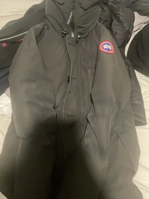 Svart dunjacka från Canada Goose - Säljer en svart dunjacka från Canada Goose med huva och klassisk logotyp på bröstet. Jackan har dragkedja, knappar och är fodrad med dun för extra värme. Perfekt för kalla vinterdagar och har en stilren, lång modell.