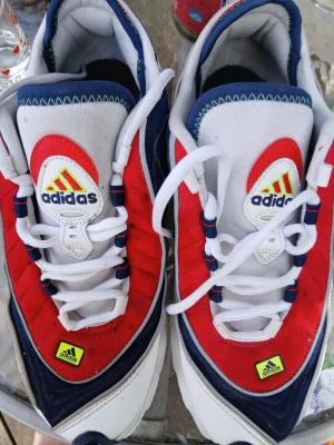 Adidas sneakers i rött, vitt och blått - Snygga Adidas sneakers med en sportig vibe i rött, vitt och blått. Ovandelen är i textil och syntet med tydliga Adidas-loggor på plösen och framtill. Sulan har coola detaljer i svart, rött och gult. Skorna har vita snören och låg profil för en klassisk look.