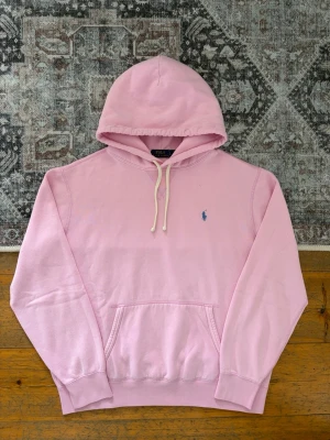 Ljusrosa Polo Ralph Lauren Fleece Hoodie - Storlek L - Hej! 👋 Säljer en riktigt snygg ljusrosa Polo Ralph Lauren hoodie i storlek L med mörkblå pony-logga. 🌸🐎 Tröjan är i mycket gott skick och känns superfräsch. Det finns 4 st minimala prickar på ena ärmen som knappt syns vid användning (se sista bilderna). 100% äkta med originaltvättlappar kvar och en fullt fungerande QR-kod för superenkel verifiering ✅ Perfekt färgklick till garderoben! Fraktar snabbt! 3💬 🚛📦✨ 