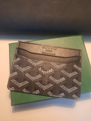 Svart kortfodral  - Snyggt svart kortfodral från med det klassiska vita och gråa mönstret. Tillverkat i läder med flera fack för kort. Märkets logga syns tydligt framtill. Perfekt för dig som vill ha en stilren och exklusiv accessoar. Pris går att diskuteras! JÄTTEBRA KVALITE!