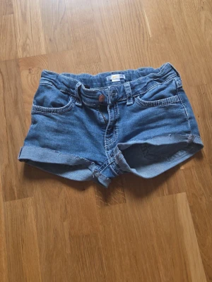 Blå jeansshorts med fickdetaljer - Snygga blå jeansshorts med uppvikta ben och dekorativa bakfickor med knappar. Klassisk femficksmodell och bälteshällor. Perfekta för varma dagar och enkel att matcha med olika toppar. OBS klippte de själv från ett par jeans.