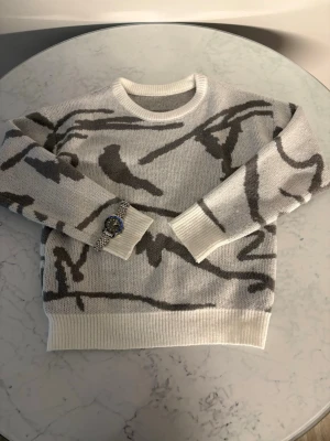 Vit och grå stickad tröja med mönster - Säljer en snygg stickad tröja i vitt med grått abstrakt mönster. Tröjan har rund halsringning, ribbade muddar och nederkant. Perfekt för dig som gillar en stilren men ändå unik look.
