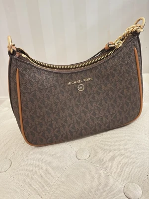 Michael Kors brun axelremsväska - Snygg brun handväska från Michael Kors (Äkta)  med klassiskt MK-mönster, guldfärgade detaljer och kedja. Väskan har dragkedja upptill och ljusbruna kanter. Perfekt för dig som gillar stilrena accessoarer med lyxig känsla.