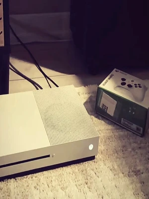 Xbox One S vit spelkonsol - Säljer en stilren Xbox One S i vit färg från Microsoft. Konsolen har en slimmad, rektangulär design med ventilerad ovansida och matta ytor. Perfekt för gaming och streaming. Original Xbox-kontrollens kartong syns också på bilden.