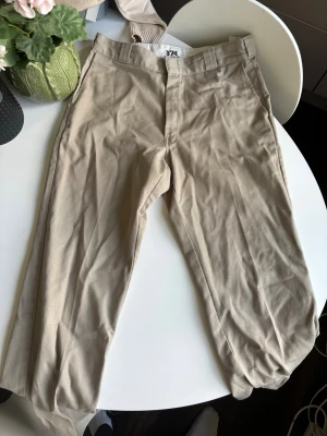 Beige Dickies 874 chinos - Säljer ett par klassiska beige Dickies 874 chinos med raka ben och normal midja. Byxorna har bälteshällor, två sidofickor och två bakfickor. Tillverkade i slitstarkt bomullsmaterial med en clean och tidlös look.