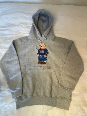 Polo Bear Ralph Lauren S - Snygg grå hoodie från Ralph Lauren med Polo Bear-tryck framtill. Klassisk huva med dragsko och stor magficka. Mjuk bomullskänsla och avslappnad passform. Perfekt för en chill och stilren look. Storlek Small.
