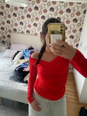 Röd offshoulder topp med rynk - Snygg röd offshoulder topp med långa ärmar och rynkade detaljer längs sidorna och ärmarna. Toppen har en figurnära passform och är tillverkad i ett stretchigt syntetmaterial som sitter bekvämt på kroppen. Perfekt för dig som vill sticka ut med en färgstark look.
