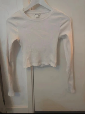 Vit ribbad croppad långärmad topp - Säljer en vit croppad långärmad topp från Monki i fint skick. Toppen är ribbad och har en rund halsringning. Perfekt att styla med höga jeans eller kjol för en clean look.