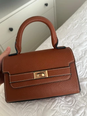 Brun läderhandväska med gulddetalj - Säljer en snygg brun handväska i läder med struktur och vit kontrastsöm. Väskan har ett stadigt handtag och en elegant guldfärgad låsdetalj på framsidan. Perfekt för dig som gillar klassisk och stilren design.