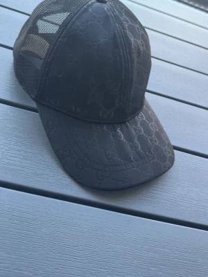 Svart keps från Gucci med mesh - Snygg svart keps från Gucci med klassiskt GG-mönster på framsidan och böjd skärm. Baksidan är i mesh för extra ventilation och har en liten metallbricka med Gucci-logga. Perfekt accessoar för en trendig streetstyle-look.