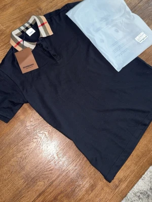 Svart Burberry t-shirt med rutig krage - Snygg svart t-shirt från Burberry med klassisk rutmönstrad krage i beige, rött och svart. T-shirten har korta ärmar och knäppning framtill. Perfekt för dig som vill ha en stilren look med en touch av lyx.