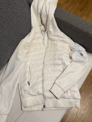 Vit cardigan från Moncler - Snygg vit pufferjacka från Moncler med huva och dragkedja framtill. Jackan har quiltad front, två fickor med dragkedja och en ikonisk Moncler-logga på ärmfickan. Mjuk bomullskänsla i ärmar och rygg, perfekt för kalla dagar.
