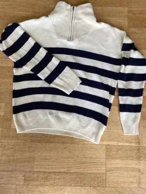 Randig stickad tröja med halvzip - Vit och marinblå stickad tröja med breda horisontella ränder och halvzip i halsen. Tröjan har lång ärm och ribbstickade muddar. Perfekt för en avslappnad och trendig look.