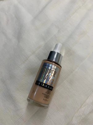 Maybelline Super Stay Skin Tint Foundation - Maybelline Super Stay 24H Skin Tint foundation i en ljus beige nyans. Kommer i en glasflaska med silvrig pipett för enkel applicering. Ger en naturlig finish och innehåller vitamin C för extra lyster. Perfekt för dig som vill ha en lätt men täckande bas.