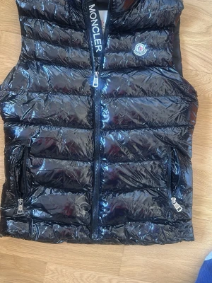 Svart glansig väst  - Snygg svart dunväst med glansigt material. Västen har två dragkedjeförsedda fickor och en hög krage. Perfekt för lager-på-lager och streetwear-vibe. Använd 1 gång! Skriv vid funderingar