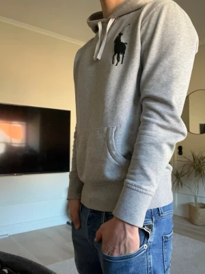 Grå hoodie från Polo Ralph Lauren - Säljer en grå hoodie från Polo Ralph Lauren med stor svart logga på bröstet. Hoodien har huva med vita snören och en klassisk magficka. Tillverkad i mjukt sweatshirtmaterial som är riktigt skönt. Perfekt till jeans eller joggers.