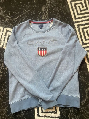 Blå sweatshirt från GANT - Ljusblå sweatshirt från GANT med broderad logga och amerikanskinspirerat sköldmärke på bröstet. Tröjan har rund halsringning, långa ärmar och ribbade muddar vid ärmslut och nederkant. Perfekt för en avslappnad och stilren look.