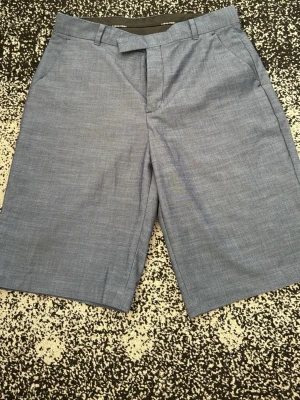 Blåa klassiska shorts Calvin Klein - Säljer ett par stilrena blåa shorts från Calvin Klein med normal passform. Shortsen har bälteshällor, sidofickor och en bakficka med knapp. Perfekta för sommaren och tillverkade i ett lätt bomullsmaterial.