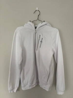 Vit hoodie från Sail Racing - Snygg vit hoodie från Sail Racing med dragkedja framtill och en extra bröstficka med logga. Hoodien har huva, långa ärmar och ribbade muddar. Perfekt för dig som gillar stilren och sportig design. Materialet känns mjukt och bekvämt.