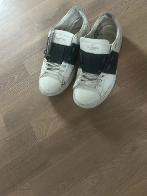 Valentino vita sneakers med mörkblå  detalj - Säljer ett par snygga vita sneakers från Valentino med en bred blå rem över mitten. Skorna har klassisk låg modell, vita snören och är tillverkade i läder. Perfekta för dig som gillar stilrena och exklusiva sneakers med en twist.
