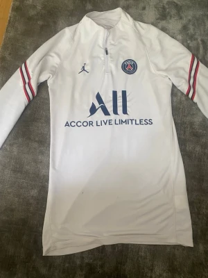 Vit PSG träningströja Jordan - Säljer en vit långärmad träningströja från PSG och Jordan med halv dragkedja. Tröjan har blå och röda detaljer på ärmarna, PSG-logga på bröstet och sponsortryck framtill. Perfekt för fotbollsträning eller chill. Materialet är troligtvis polyester.