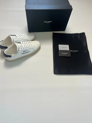 Saint Laurent vita sneakers med svart detalj storlek 42 - Snygga vita sneakers från YSL med svart detalj på hälen och handskriven logga på sidan. Skorna har klassisk låg profil, vita skosnören och gummisula med Saint Laurent Paris-tryck. Tillverkade i läder för en lyxig känsla. Skorna har storlek 42 och är aldrig burna och helt nya