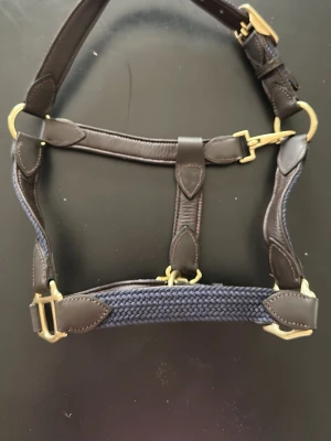 Marinblå grimma med mässingsdetaljer - Snygg grimma i marinblått flätat tyg med detaljer i mörkbrunt läder och guldiga spännen. Grimman har justerbara remmar för perfekt passform. Köpt på RiderSport i Båstad!
