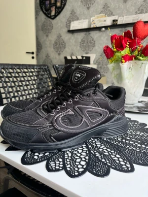 Svarta Dior B30 sneakers - Svarta Dior B30 sneakers med tydliga vita kontrastsömmar och stora reflekterande CD-loggor på sidorna. Ovandelen är i mesh och syntetmaterial, med robust sula och snörning framtill. Märkesdetaljer på plös och häl ger en exklusiv vibe.