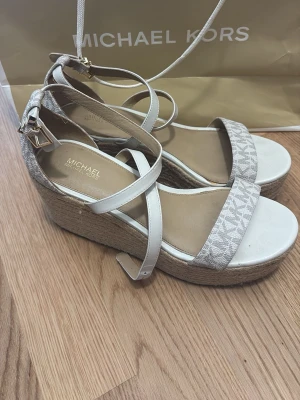 Michael Kors vita kilklackssandaler - Snygga vita sandaler från Michael Kors med kilklack i flätad jute och remmar i läder. Främre remmen har MK-logga i grått och spänne i guld. Perfekta för sommaren med sin trendiga och bekväma design.