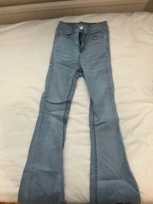Ljusblå utsvängda jeans - Säljer ett par ljusblå jeans med utsvängda ben och klassisk femficksdesign. Jeansen har hög midja och stängs med knapp och dragkedja. Perfekta för dig som gillar retrostil.