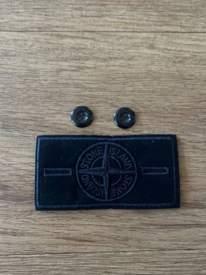 Svart Stone Island patch med knappar - Säljer en helt ny all black patch från Stone Island med tillhörande två svarta knappar. Perfekt om du tappat bort eller blivit av med din patch och vill sätta dit en ny patch på jackan, hoodien eller tröjan.  PRIS KAN DISKUTERAS!!!